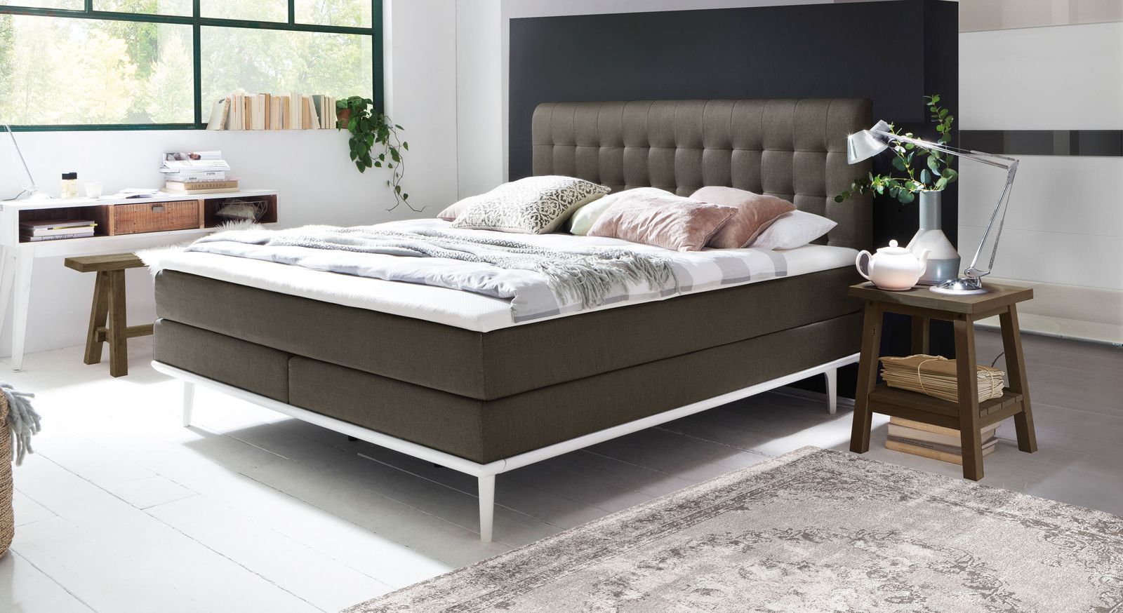 Moderne Farbkombination: das Boxspringbett "Tromello" mit einem anthrazitfarbenen Bezug und weißen Füßen sowie Unterleisten.