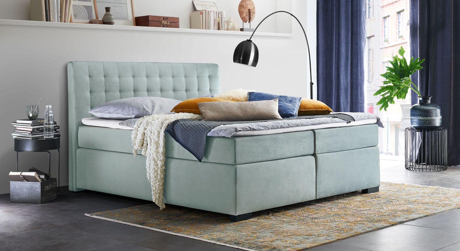 Das klassisch-elegante Bettkasten-Boxspringbett "Edessa" &ndash; hier in eisblauem Velours-Webstoff mit graphitfarbenen Holzf&uuml;&szlig;en.