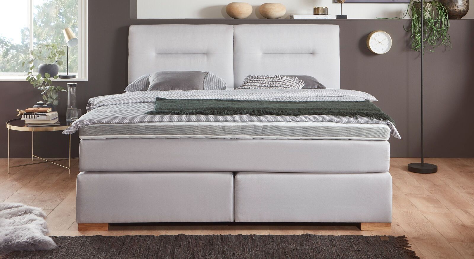 Das moderne Boxspringbett "Carlita" &ndash; hier mit hellgrauem Webstoff-Bezug und eichefarbigen Holzf&uuml;&szlig;en.