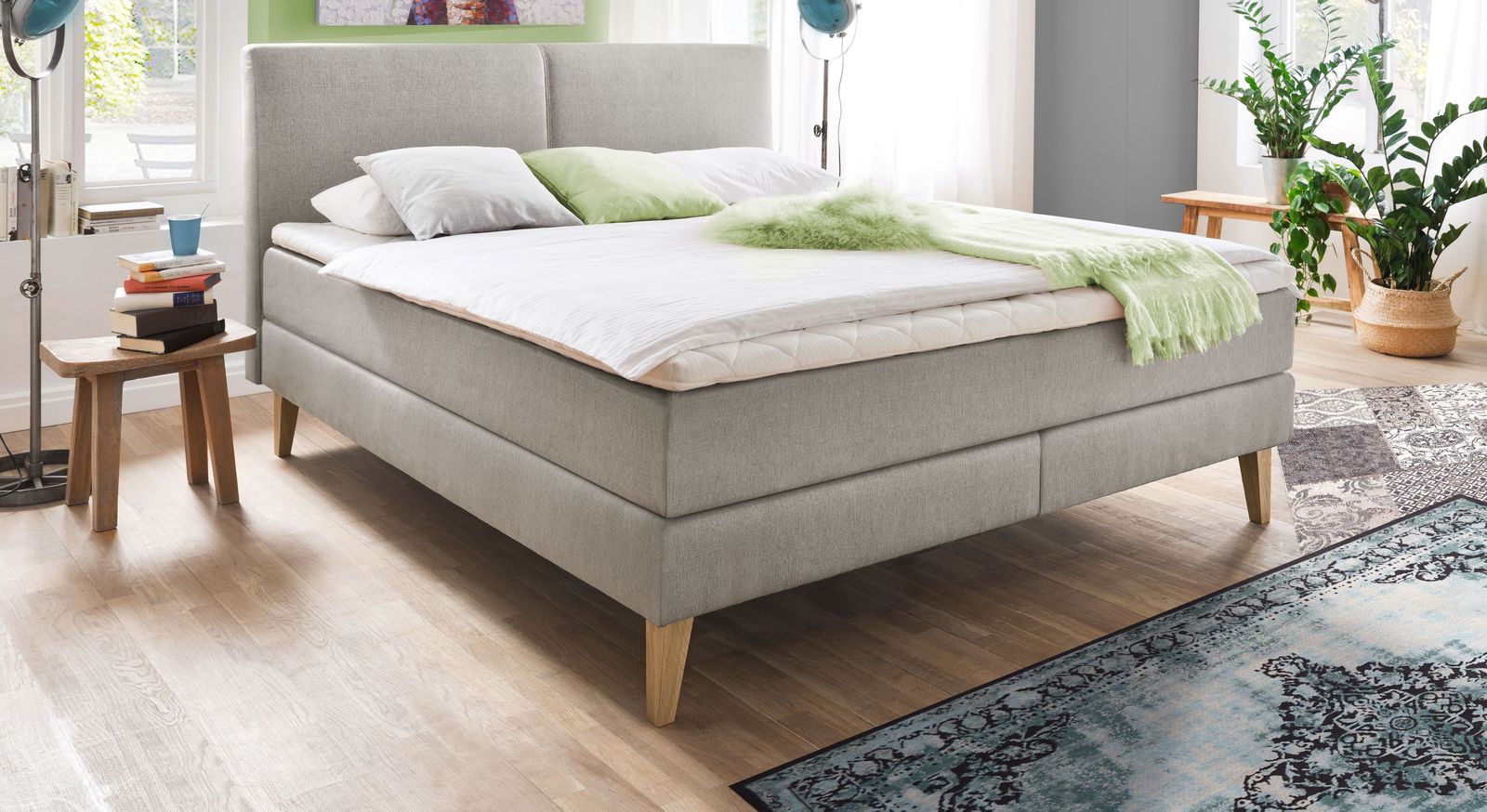 Gem&uuml;tliches skandinavisches Design: das Boxspringbett "Jaramillo" mit ausgestellten Massivholzf&uuml;&szlig;en und hellgrauem Webstoffbezug.