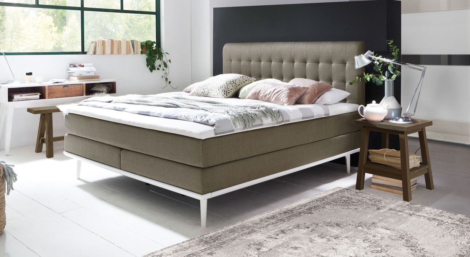 Gem&uuml;tlichkeit pur: das Boxspringbett "Tromello" &ndash; hier mit einem taupefarbenen Webstoff inklusive wei&szlig;en Unterleisten und F&uuml;&szlig;en aus Massivholz.